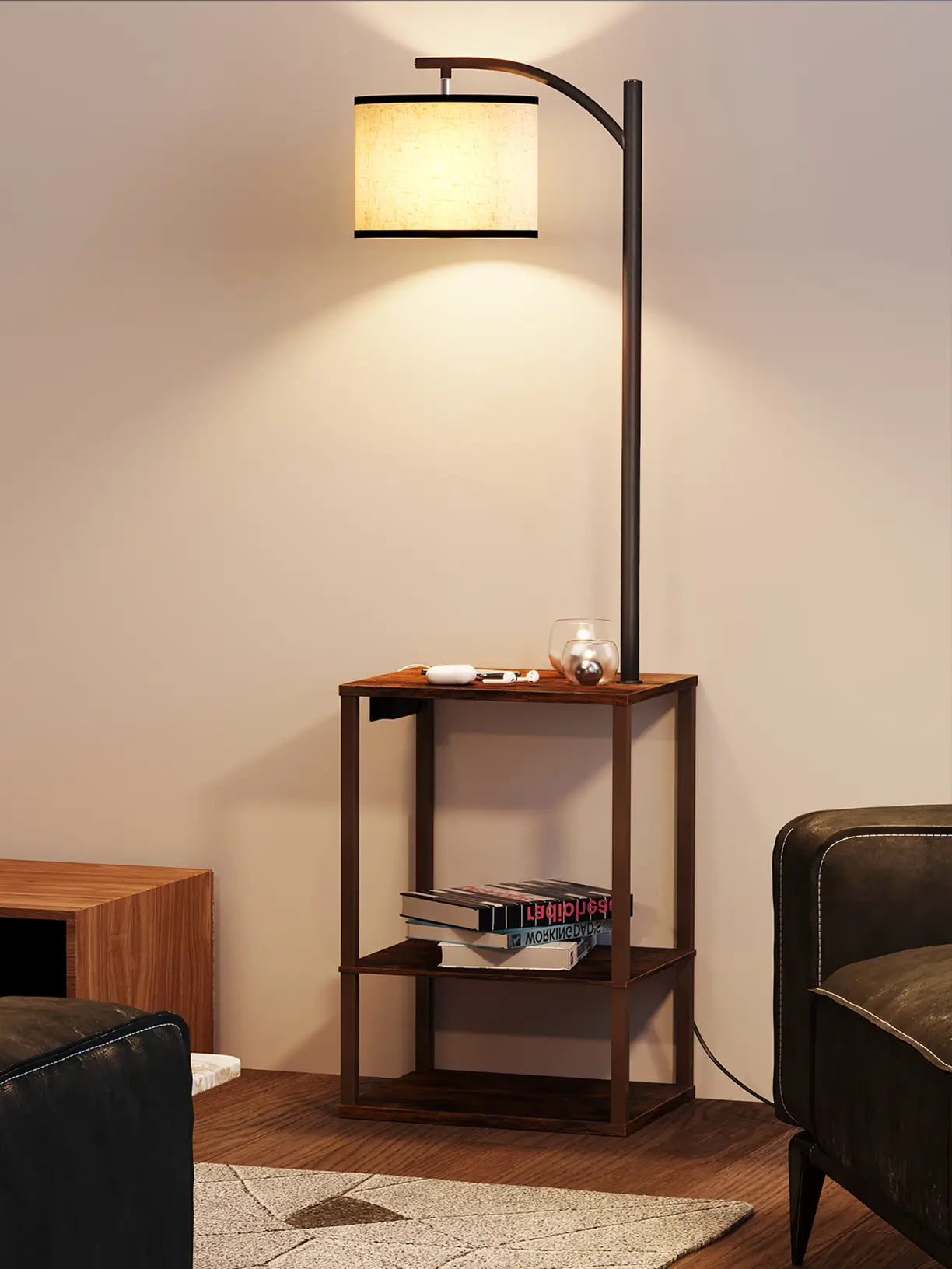 An industrial-style floor lamp with a dark metal frame and a light fabric shade, incorporating a two-tier side table for books and essentials, perfect for a cozy living space next to a leather armchair.