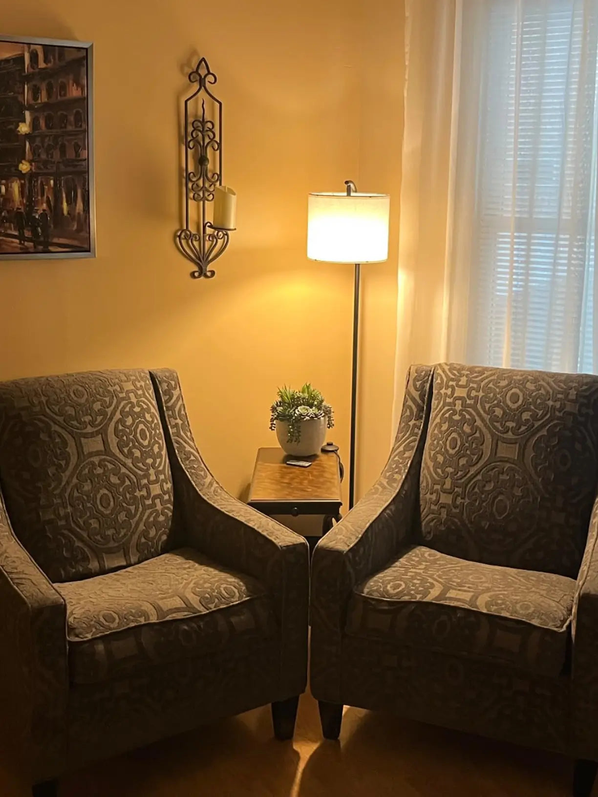 A sleek, modern arched floor lamp with a white fabric drum shade, extending over a plush gray sectional sofa in a bright living room, providing ambient overhead lighting.