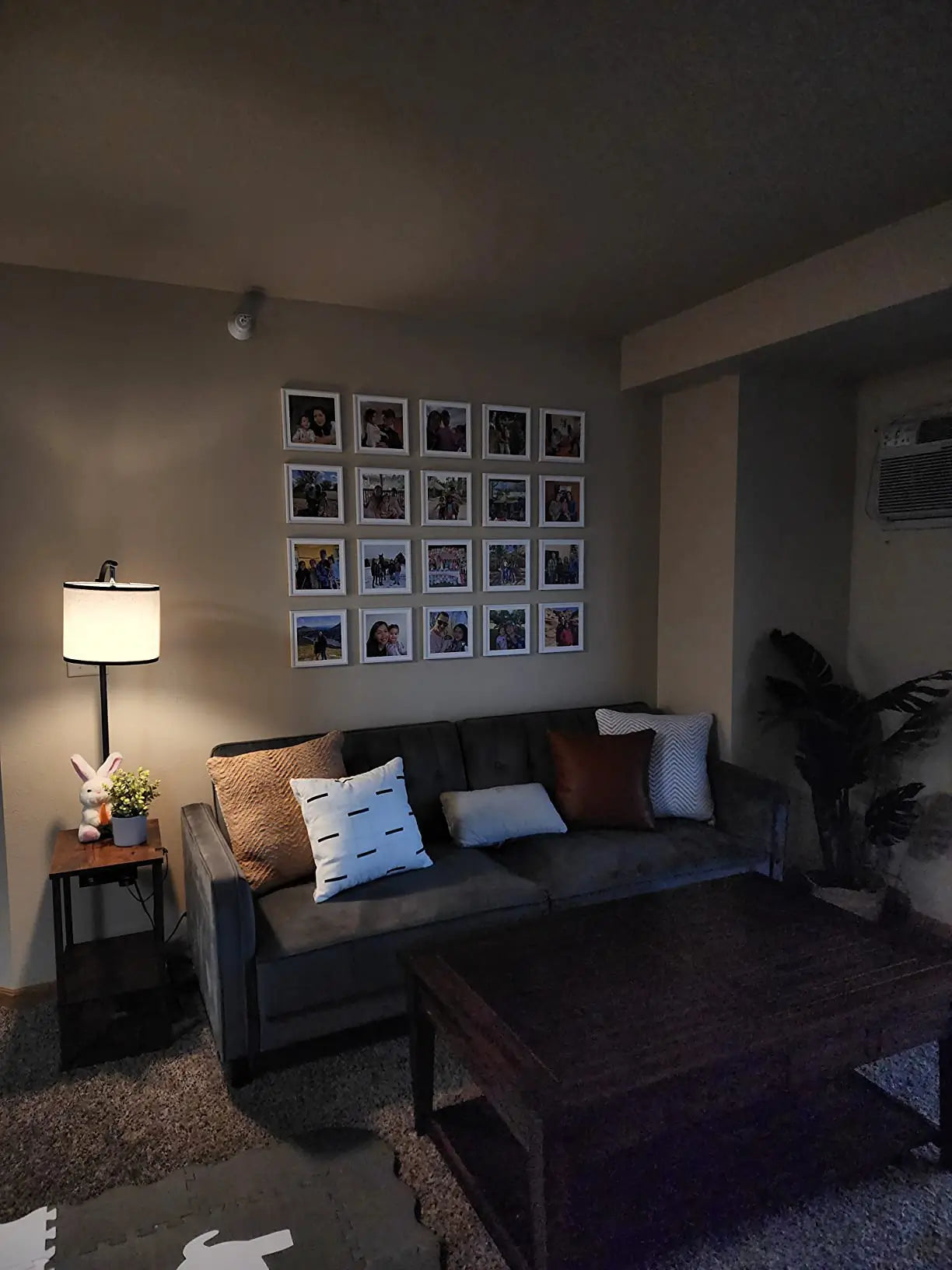 A contemporary floor lamp with a dark base and light shade, standing next to a small side table, illuminating a living room with a grey sofa and a gallery wall of framed photos.